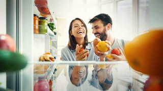 Una pareja sonriente abre el refrigerador para buscar unas frutas