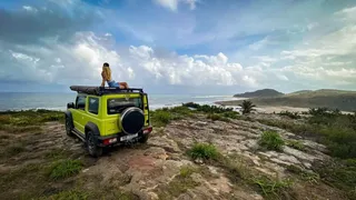 Suzuki Jimny 2026 color Kinetic Yellow estacionado en terreno de tierra con fondo de playa y montañas