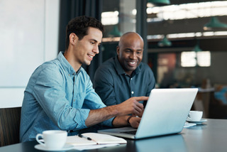 Dois homens utilizam um notebook enquanto tomam um café.