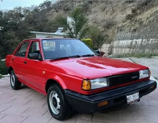 Auto Nissan Tsuru rojo clásico en excelente estado, estacionado en entrada de adoquín con fondo de vegetación y cerro, vista frontal lateral