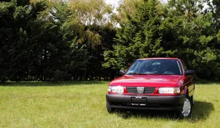 Auto rojo estacionado en campo verde con árboles detrás