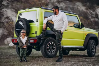 Suzuki Jimny amarillo vibrante estacionado sobre terreno de grava en entorno natural, con padre e hija apoyados junto al vehículo.