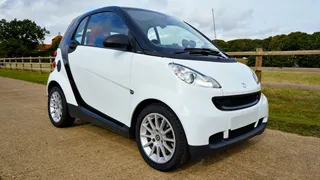 Vista lateral de un Smart Fortwo blanco con célula de seguridad negra, estacionado en un camino rural junto a una cerca de madera