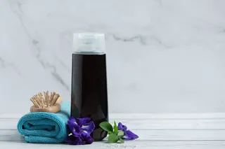 shampoo violeta rodeado de flores, junto a un cepillo y toalla sobre mesa
