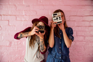 Mulheres utilizam câmera analógica na frente de uma parede rosa