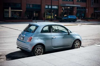 Fiat 500 azul estacionado en una calle con un edificio de ladrillo de fondo.