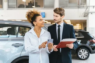 Dos personas en una concesionaria analizan opciones de financiamiento desde un celular mientras revisan documentación, con un auto compacto exhibido detrás en el salón de ventas