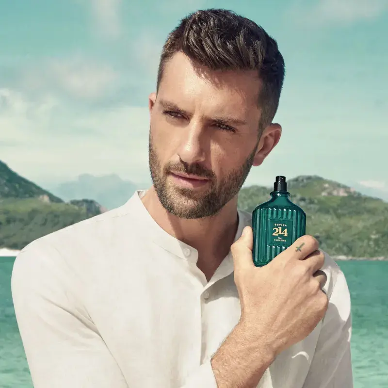Homem de camisa branca segurando o perfume Botica 214 Fiji Paradise com paisagem litorânea ao fundo