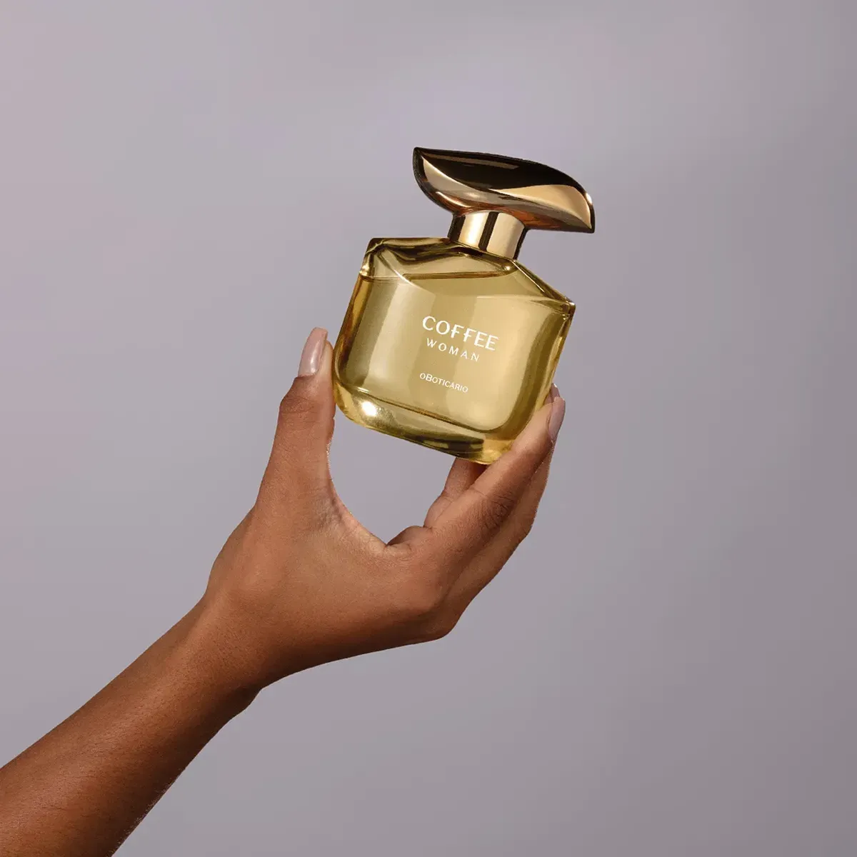 Mão feminina segurando o frasco do perfume O Boticário Coffee Woman contra fundo neutro