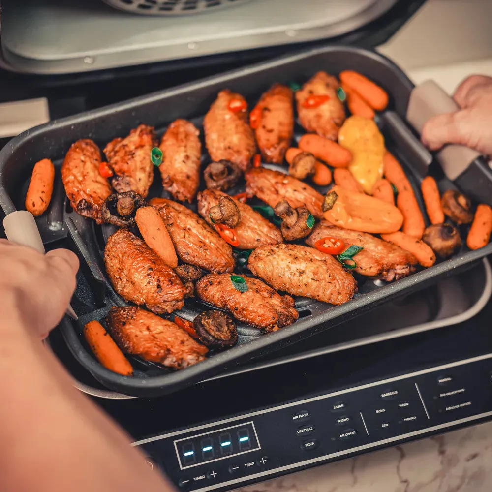 Close-up de mãos removendo a bandeja da Fritadeira Elétrica Air Fryer Wap Barbecue Heat, repleta de asinhas de frango crocantes, cenouras e cogumelos assados.