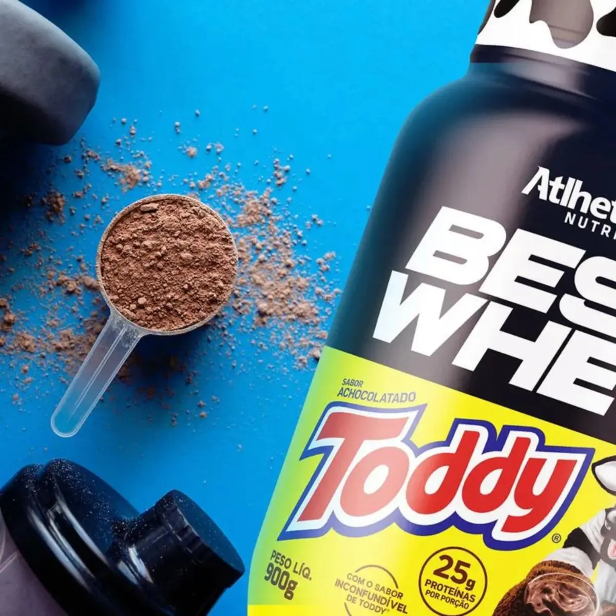 Colher medidora cheia de pó de chocolate do Whey Best Whey Toddy ao lado do pote, em um fundo azul, com respingos de pó.