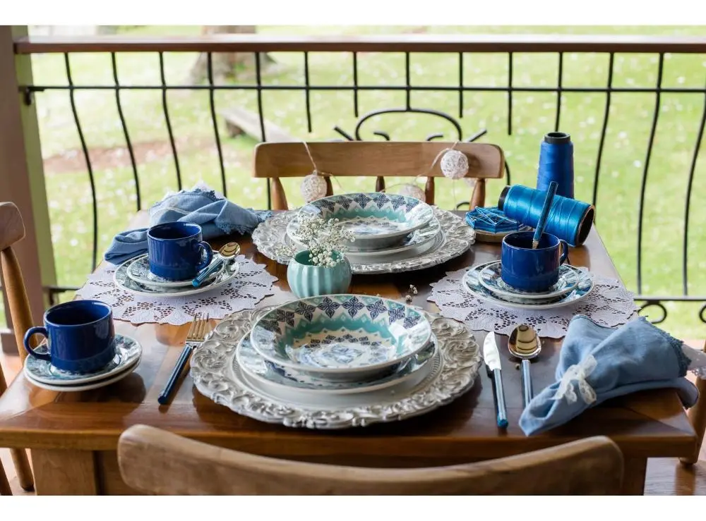 Mesa posta com aparelho de jantar Oxford Cerâmica Lola, incluindo pratos, bowls, xícaras e guardanapos azuis, dispostos em uma varanda.