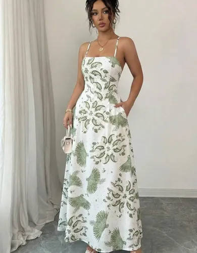 Mulher usando vestido longo branco com estampa verde de folhas e pássaros, alças finas, com as mãos nos bolsos.