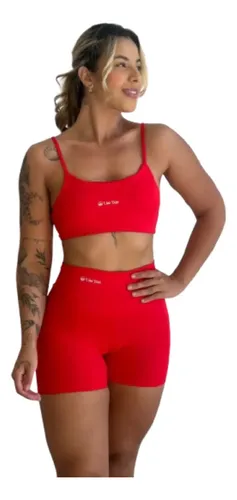 Modelo feminina sorrindo, posando frontalmente com um conjunto fitness vermelho, composto por top de alça fina e shorts meia coxa de cintura alta, em fundo branco isolado.