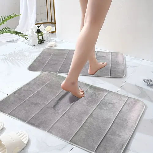 Pernas de uma mulher pisando em tapetes cinza no banheiro, com azulejos brancos e itens de higiene pessoal ao fundo.