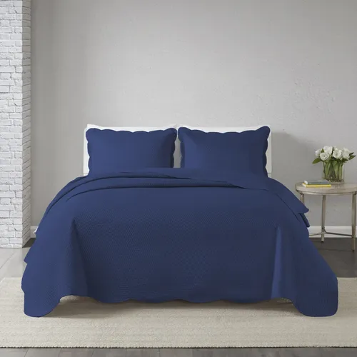 Cobre leito de casal azul marinho com textura matelassê e acabamento em bordas onduladas, acompanhado de dois porta-travesseiros coordenados em um quarto com parede de tijolos brancos.