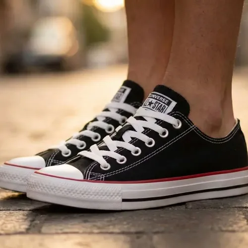 Pés usando tênis Converse Chuck Taylor All Star pretos com cadarços brancos e detalhe vermelho na lateral.