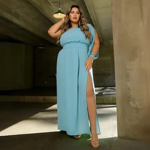 Modelo plus size usando vestido longo azul claro de um ombro só com fenda lateral em ambiente de concreto.