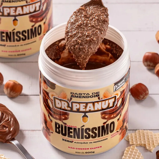 Pote de 600g de pasta de amendoim Dr. Peanut sabor Bueníssimo com uma colher retirando uma porção cremosa do produto, cercado por avelãs e pedaços de wafer.