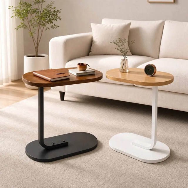 Duas mesas de apoio lateral em formato C, uma com base e estrutura preta e outra com base e estrutura branca, ambas com tampos em tons de madeira sobre um tapete em frente a um sofá claro.