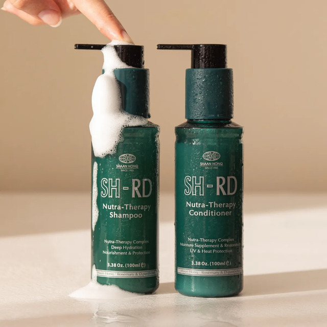 Frascos de Shampoo e Condicionador SH-RD Nutra-Therapy de 100ml posicionados lado a lado, com uma mão pressionando o pump do shampoo e criando espuma branca sobre um fundo bege.