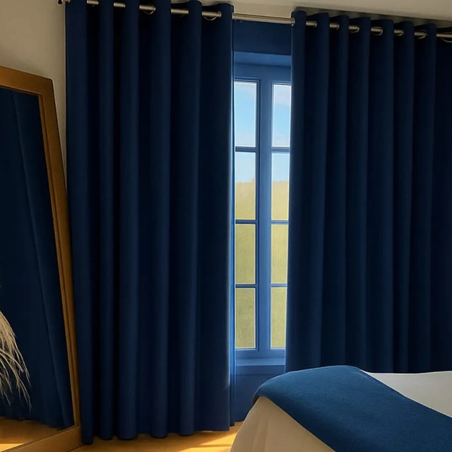 Cortina blackout de tecido azul marinho instalada em uma janela, acompanhada por um espelho com moldura de madeira e um vaso decorativo com plantas secas ao lado de uma cama.
