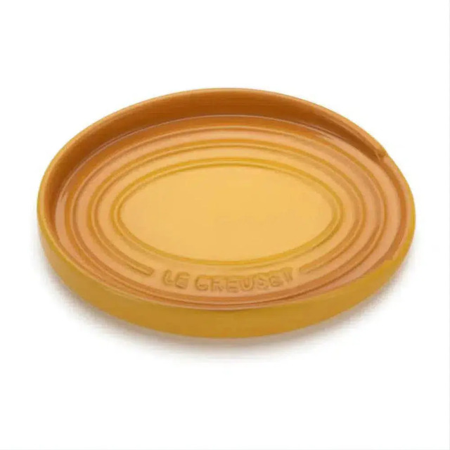 Descanso de colher oval da marca Le Creuset na cor amarelo mostarda, feito de cerâmica com relevo do nome da marca no centro