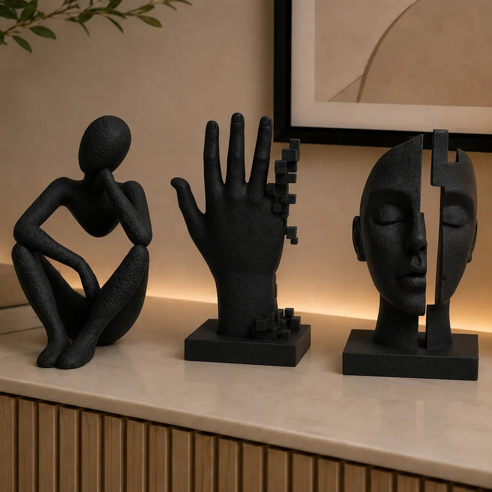 Trio de esculturas decorativas pretas sobre um aparador, incluindo uma figura humana sentada, uma mão com detalhes em cubos e uma cabeça dividida ao meio.