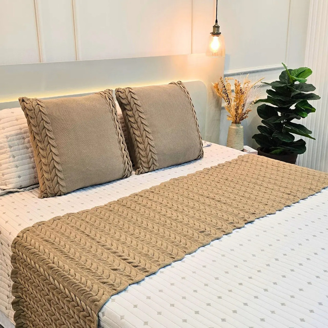 Kit de peseira e duas capas de almofada em tricot cor bege claro dispostos sobre uma cama queen com colcha branca e elementos decorativos ao fundo