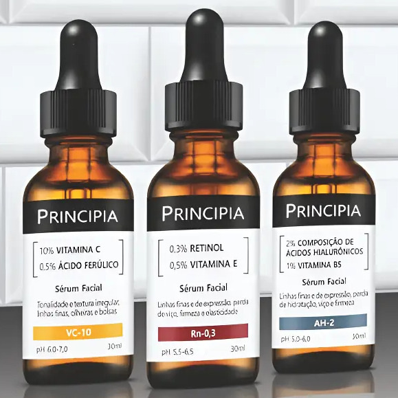 Três frascos de sérum facial da marca Principia, modelos VC-10, Rn-0,3 e AH-2, dispostos lado a lado com conta-gotas em fundo de azulejos brancos.