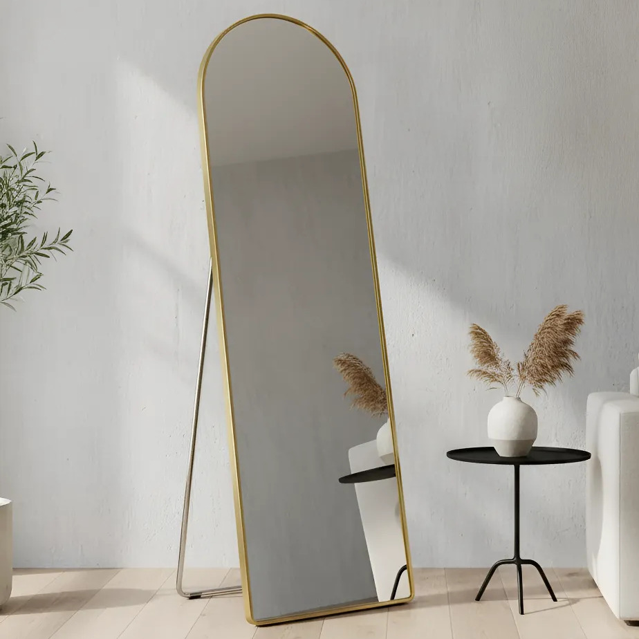 Espelho de corpo inteiro com moldura dourada arqueada, posicionado sobre suporte de chao em um ambiente clean com vaso decorativo ao lado.