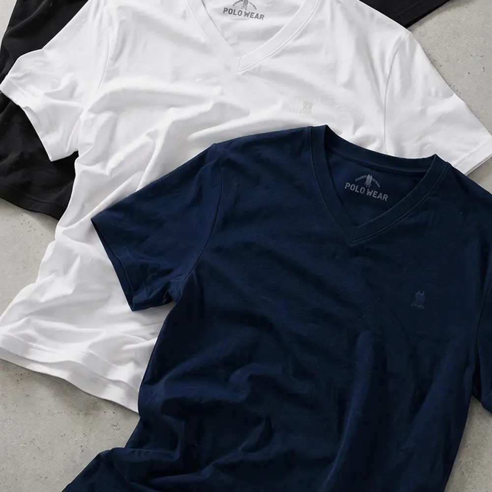 Duas camisetas básicas Polo Wear com gola V, uma na cor branca e outra na cor azul marinho, dispostas sobre uma superfície cinza.