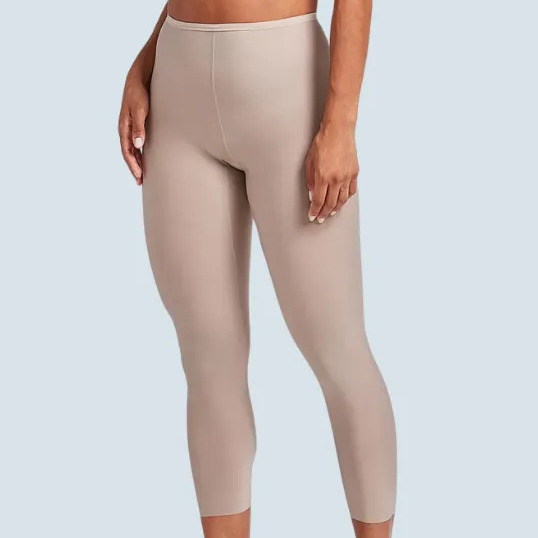 Modelo vestindo calça capri modeladora Liz cor bege, combinada com sutiã da mesma tonalidade e salto alto, em fundo azul claro.