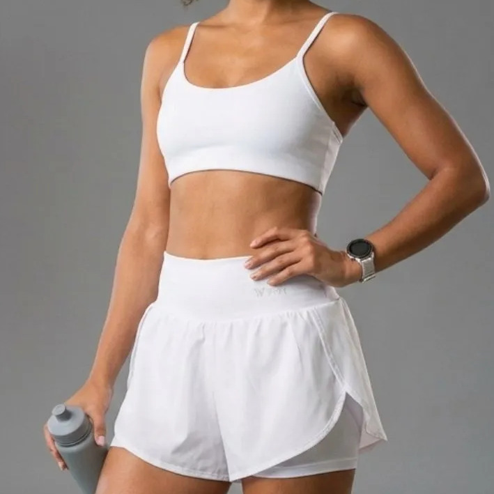Mulher vestindo conjunto fitness branco composto por top de alças finas e short duplo com garrafa de água na mão
