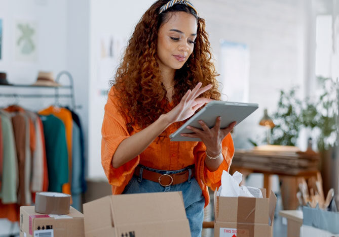 Mercado Pago: Empresária vestindo blusa laranja e calça jeans, segurando tablet pesquisando sobre o ticket médio. Há uma arara de roupas e caixas de sua loja atrás dela.