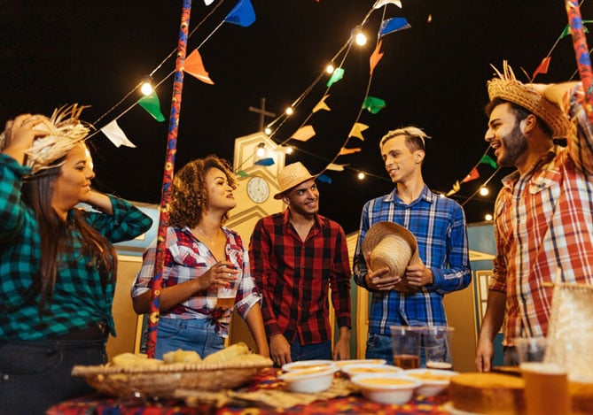 Mercado Pago: duas mulheres e três homens vestidos com camisas xadrez e chapéus de palha conversando sobre como economizar para a festa junina, ao redor de mesa com comidas típicas. Ao fundo, há uma igreja amarela e bandeirinhas penduradas.