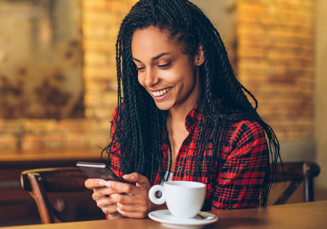 Mercado Pago: Mulher com blusa xadrez sentada no balcão de um café segurando o celular e buscando meios de como guardar dinheiro. Há uma xícara de café em sua frente.