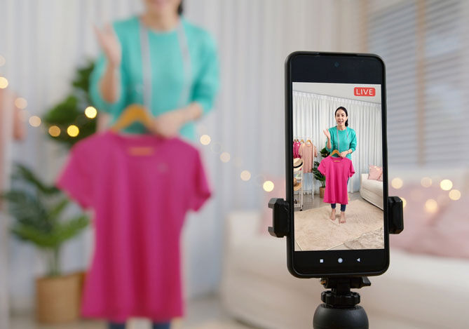 Mercado Pago: imagem de empreendedora de blusa verde segurando uma blusa rosa em frente a um celular que está gravando conteúdo para uma estratégia de vendas no Instagram.