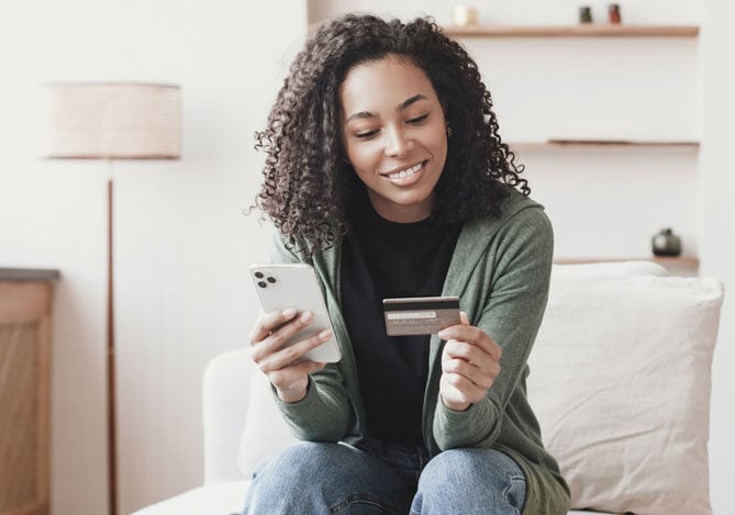 Imagem de uma mulher segurando um celular em uma das mãos e um cartão na outra para realizar um pagamento parcelado via Link de Pagamento Mercado Pago que recebeu por meio de aplicativo de mensagens.