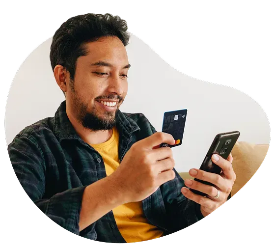 Una persona sentada cómodamente 
en su casa usa Mercado Pago desde su teléfono celular mientras sostiene 
la tarjeta Mercado Pago.