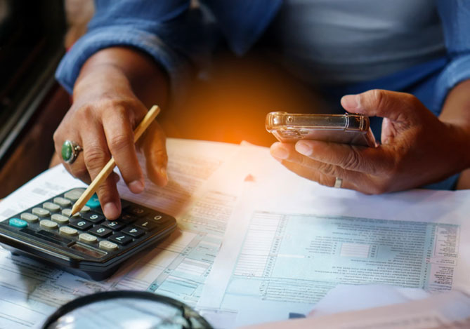 Mercado Pago: imagem das mãos de uma pessoa sobre uma mesa enquanto ela elabora um planejamento financeiro para sair do vermelho. Na imagem, é possível ver que em uma das mãos ela segura um celular onde vê algumas de suas despesas e, na outra, segura um lápis e digita em uma calculadora. Sobre a mesa, é possível ver algumas contas e faturas.