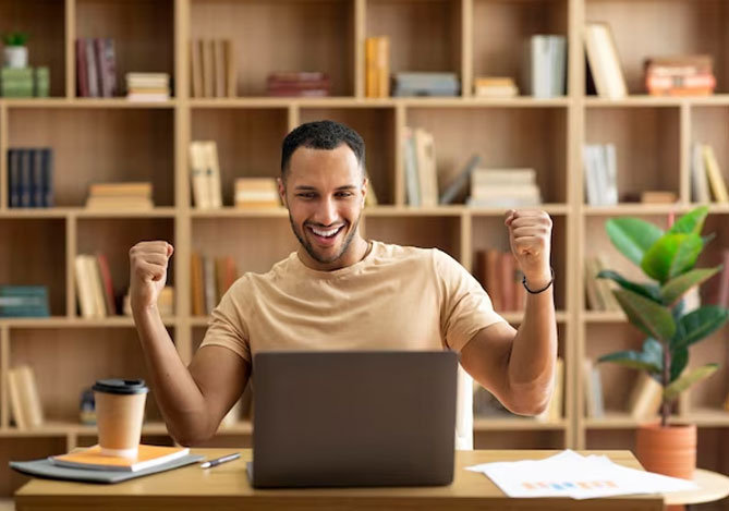Homem com camiseta na cor beige comemorando os pontos que ganhou no novo programa de recompensas Meli+ do Mercado Pago e Mercado Livre em frente ao laptop aberto em sua mesa de escritório.