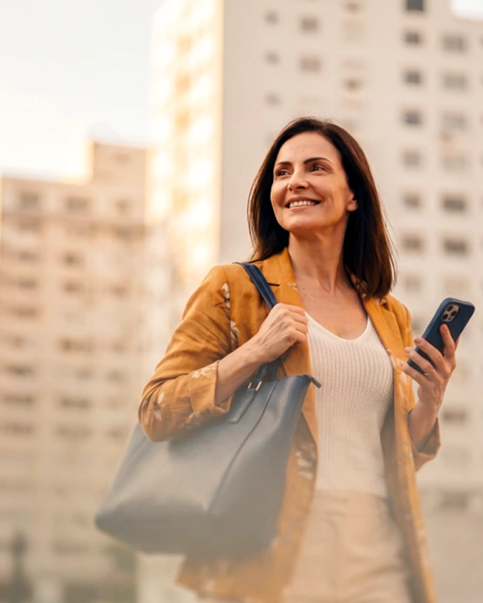 Mulher sorridente com jaqueta mostarda, bolsa no ombro e celular na mão, caminhando por uma rua da cidade