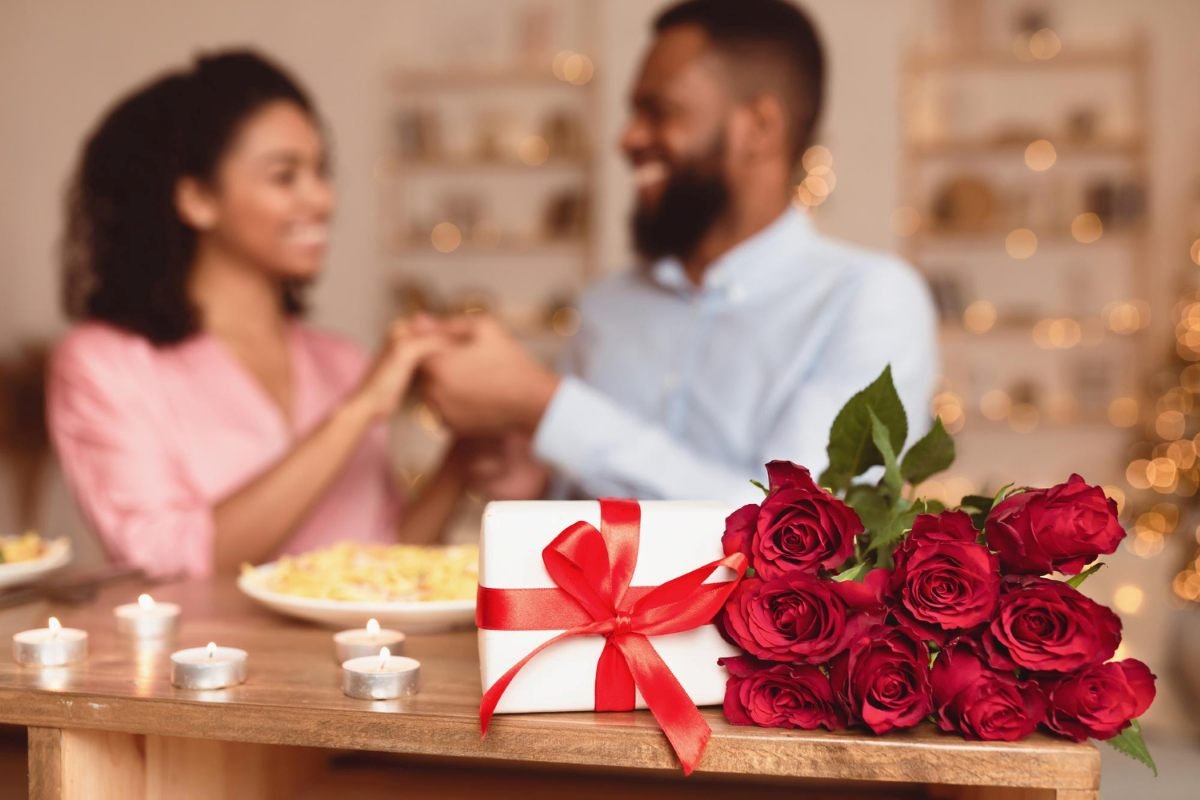 Casal sorrindo ao abrir um kit presente embalado com laço, sobre mesa com flores e velas