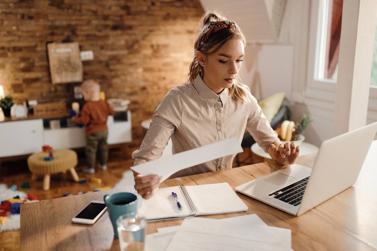 Trabalhadora autônoma sentada em mesa de trabalho home office, com notebook aberto e documentos financeiros para declara
