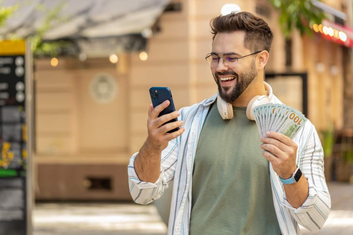Jovem olhando para o celular e  segurando notas de dólar