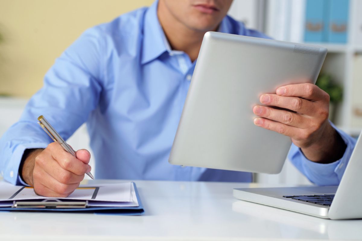 Homem faz notas em mesa com prancheta, tablet e notebook