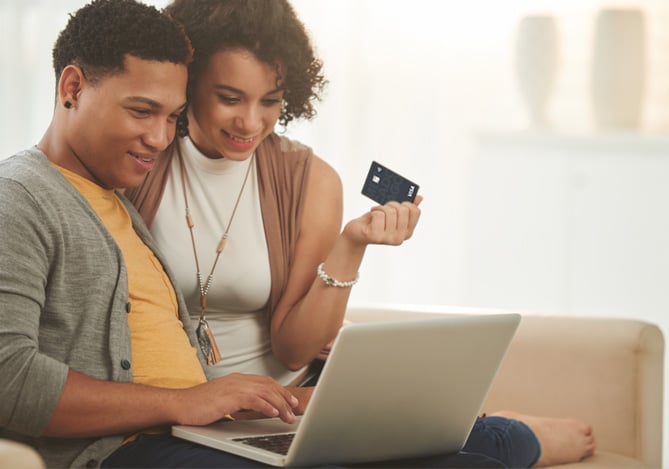 Homem mexendo no computador e mulher segurando cartão de crédito enquanto conferem a opção de parcelamento de fatura do Mercado Pago